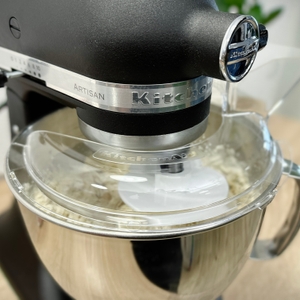 Die Küchenmaschine KitchenAid Artisan verarbeitet Teig mit einem Knethaken. Der Spritzschutzdeckel ist während des Vorgangs montiert.