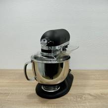 Die KitchenAid Artisan steht vollständig montiert auf einer Holzplatte. Die Schüssel ist eingesetzt, der Spritzschutz montiert.