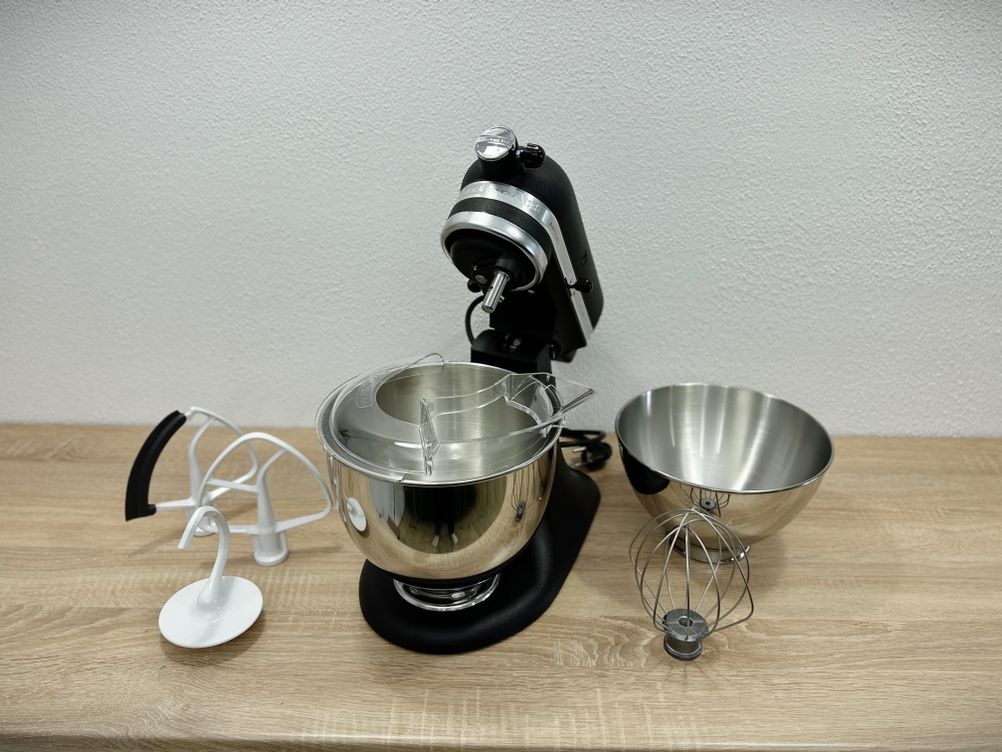 Die Küchenmaschine KitchenAid Artisan steht mit geöffnetem Motorkopf auf einem Holztisch. Rührschüssel, zweite Schüssel und alle Aufsätze liegen daneben.