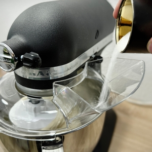 Zucker wird in die laufende Küchenmaschine KitchenAid Artisan über die Spritzschutzöffnung eingefüllt. Der schwarze Motorkopf ist deutlich erkennbar.