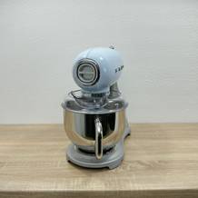 Die Küchenmaschine Smeg SMF02PBEU steht in voller Ansicht auf einer Holzarbeitsfläche. Das Gerät ist komplett montiert mit Schüssel und Spritzschutz.