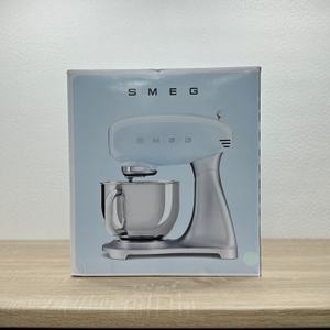 Auf der Verpackung ist die Smeg-SMF02PBEU-Küchenmaschine von der Seite abgebildet. Die Darstellung zeigt das Gerät in Silberoptik mit Schüssel.