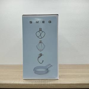Die Verpackungsseite der Smeg SMF02PBEU zeigt Abbildungen des Zubehörs: Flachrührer, Schneebesen, Knethaken und Spritzschutz. Das Logo ist oben mittig platziert.