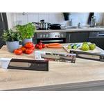 Drei verpackte Küchenmesser verschiedener Marken – Wüsthof, WMF, Zwilling – stehen nebeneinander auf einem Küchentisch mit Tomaten, Karotten, Gurken und Äpfeln.