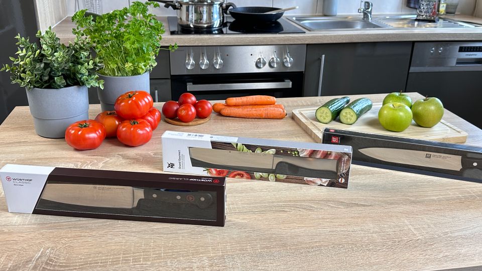 Drei verpackte Küchenmesser verschiedener Marken – Wüsthof, WMF, Zwilling – stehen nebeneinander auf einem Küchentisch mit Tomaten, Karotten, Gurken und Äpfeln.