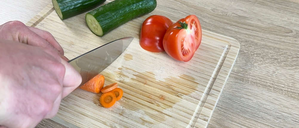 Ein WMF-Grand-Gourmet-Messer schneidet eine Karotte auf einem Schneidebrett. Im Hintergrund liegen eine Gurke und eine Tomate.
