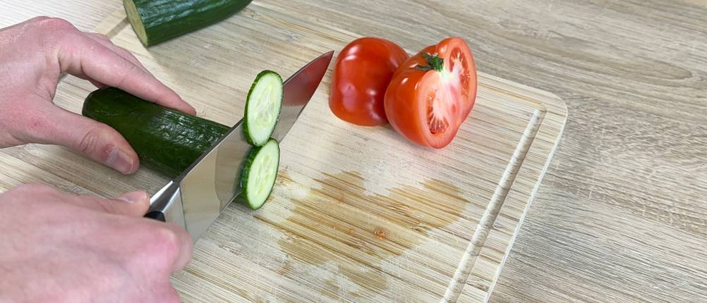 Ein Wüsthof-Chefmesser schneidet eine Salatgurke auf einem Holzbrett. Im Hintergrund liegen eine halbierte Tomate und weiteres Gemüse.