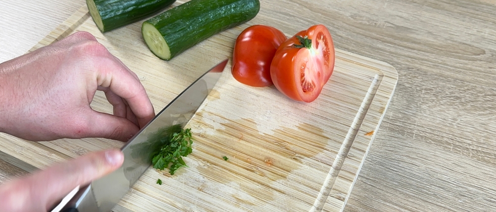 Das Wüsthof-Chefmesser zerkleinert frische Petersilie auf einem Holzbrett. Daneben liegen Gurken und Tomaten.