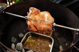 Kugelgrill mit einem Hähnchen auf dem Drehspieß und Kohlen sowie einer Fettauffangschale an Boden des Grills.