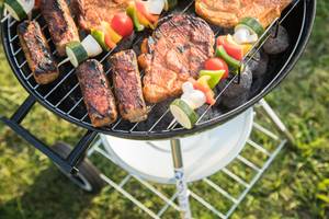Ein Kugelgrill mit Gemüsespießen, Steaks und Würstchen steht auf dem Rasen.