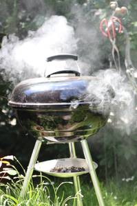 In einem Garten steht ein Kugelgrill, aus dem viel Rauch strömt.