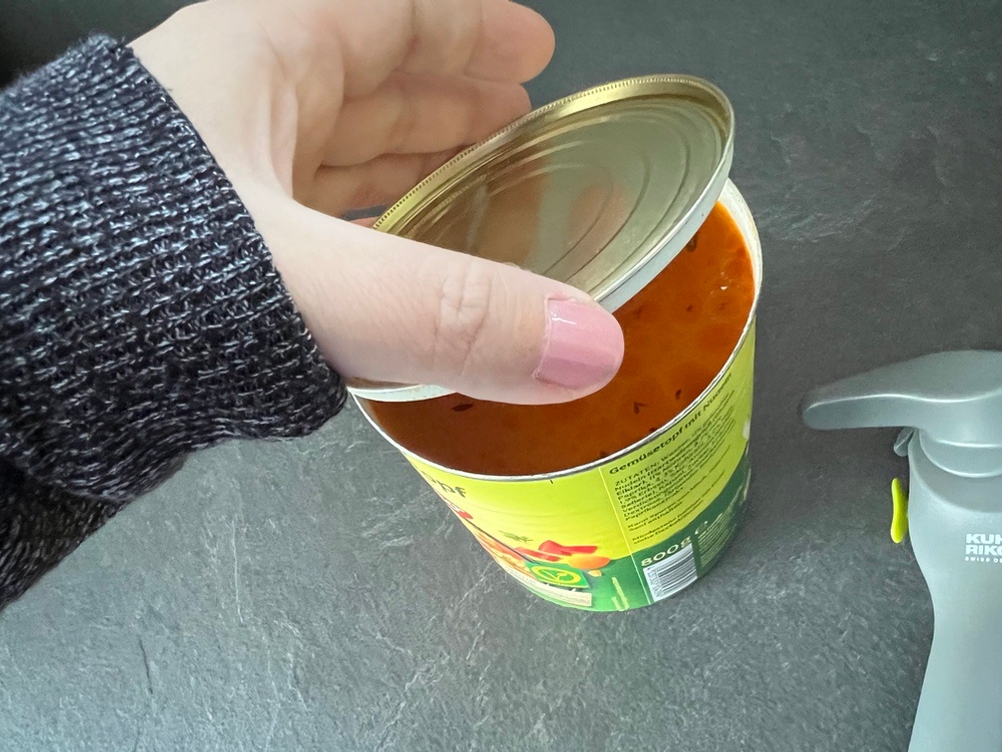 Eine Hand öffnet einen Deckel, der mit einem Dosenöffner geöffnet wurde.