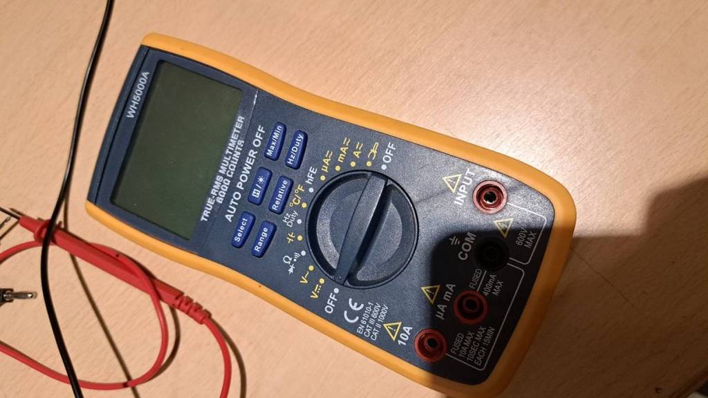 Multimeter-Test: ein digitales Messgerät in gelb-blauem Design befindet sich ausgeschaltet, inklusive rotem und schwarzem Messkabel, auf einer Holzfläche.