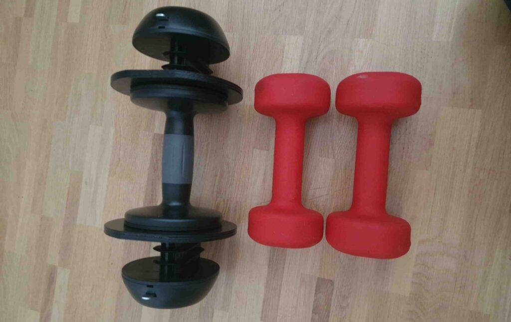 Zwei rote getestete Kurzhanteln und eine schwarze Kurzhantel mit variierbaren Gewichten liegen nebeneinander auf einem Parkettboden.