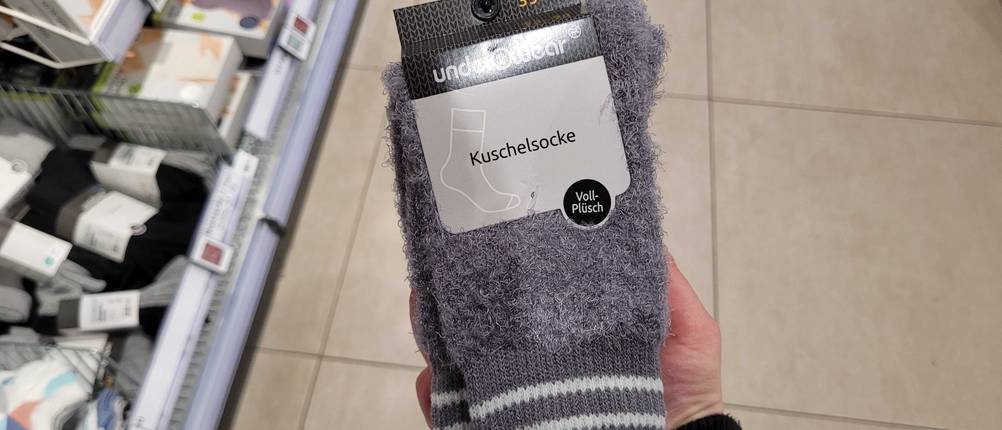 Eine Hand hält eine Packung der getesteten Kuschelsocken vor dem Einkaufsregal im Geschäft.