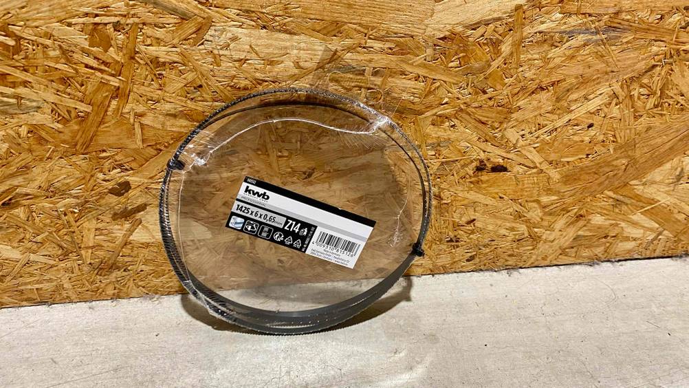 Ein getestetes Bandsägeblatt in seiner transparenten Originalverpackung vor einer Holzwand.