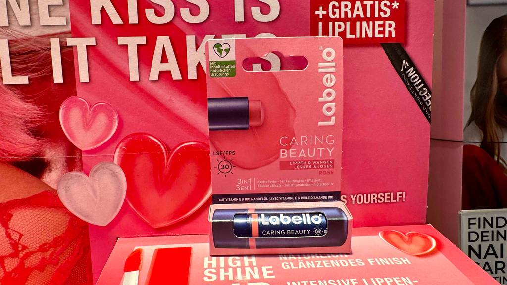 Eine getestete Lippenpflege mit Farbe steht in ihrer Verpackung vor einem Plakat.