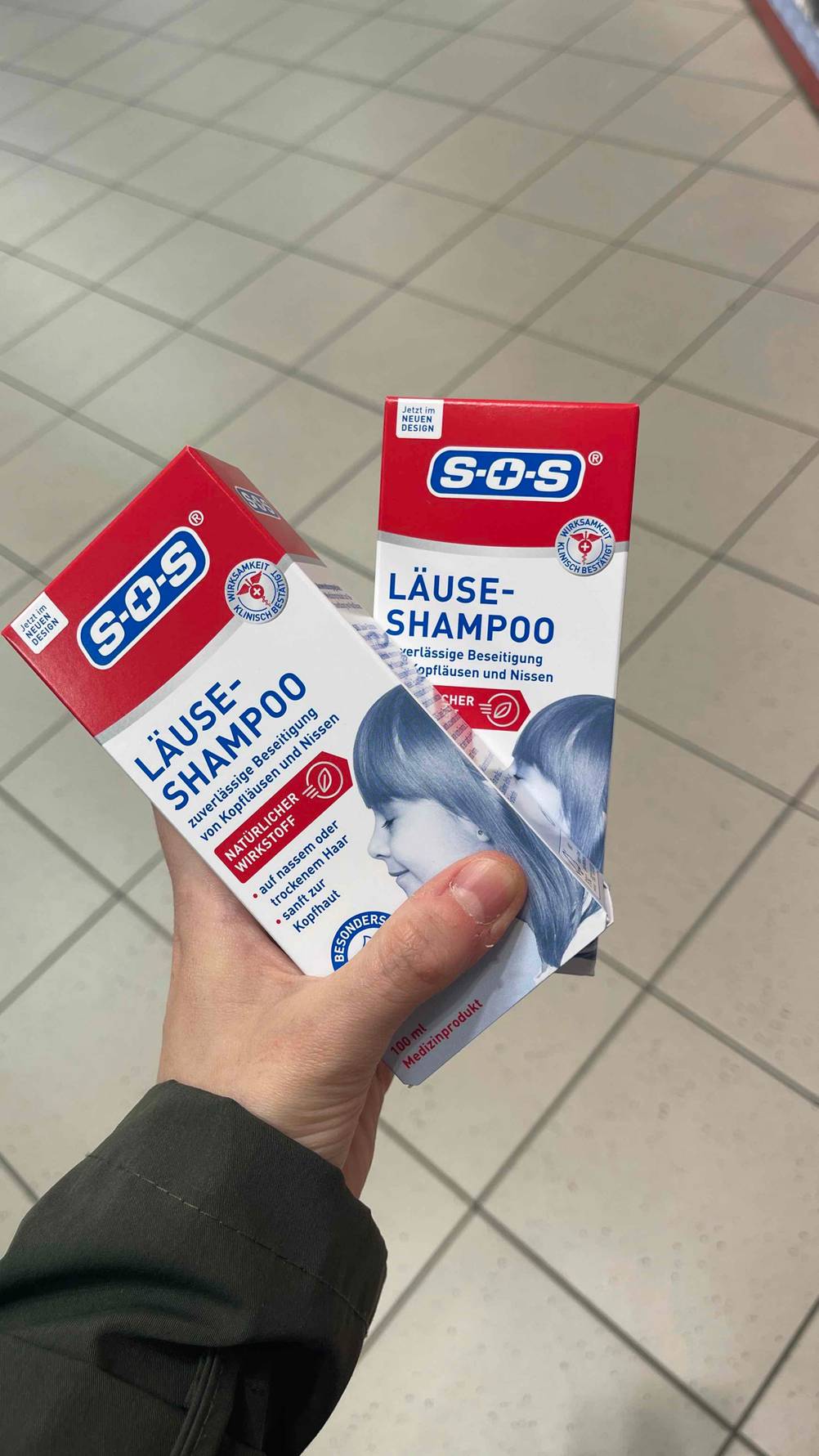 Im Test: Eine Hand hält zwei Packungen Läuse-Shampoo über einem Fliesenboden.