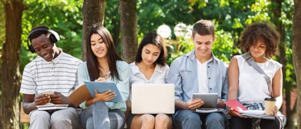Eine Gruppe junger Menschen sitzt auf der Kante einer Rasenfläche nebeneinander, alle haben ein Laptop oder ein Tablet.