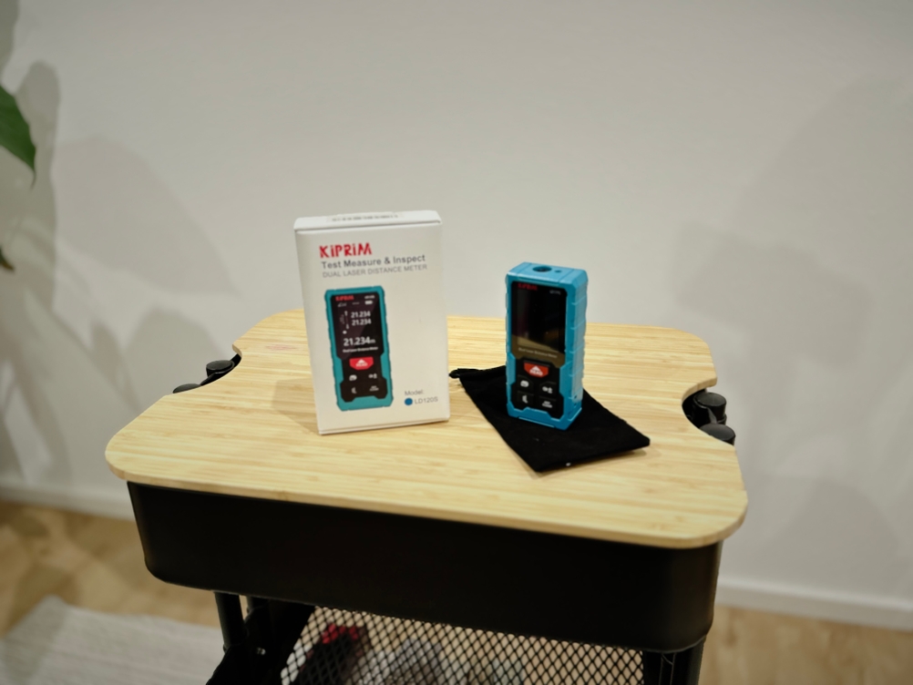 Die Verpackung sowie der Laser-Entfernungsmesser von Kiprim stehen auf einem hölzernen Untergrund vor einer weißen Wand.