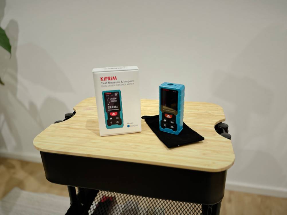 Die Verpackung sowie der Laser-Entfernungsmesser von Kiprim stehen auf einem hölzernen Untergrund vor einer weißen Wand.