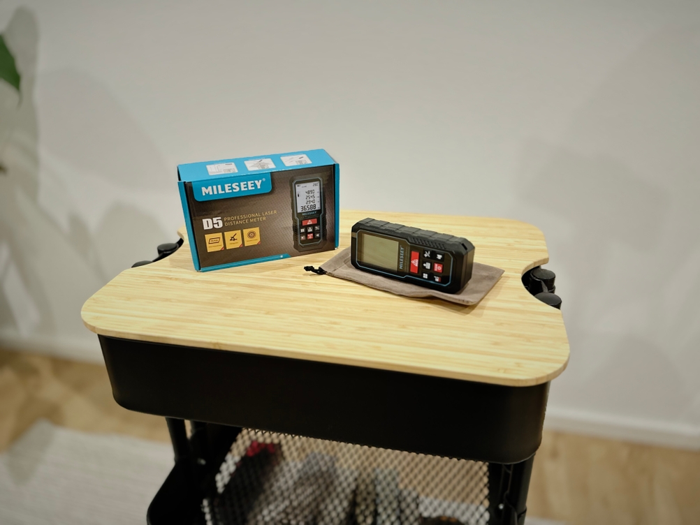 Die Verpackung sowie der Laser-Entfernungsmesser von Mileseey stehen auf einem hölzernen Untergrund vor einer weißen Wand.