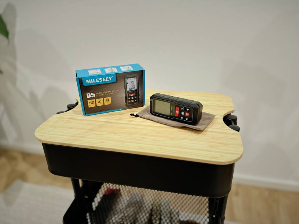 Die Verpackung sowie der Laser-Entfernungsmesser von Mileseey stehen auf einem hölzernen Untergrund vor einer weißen Wand.