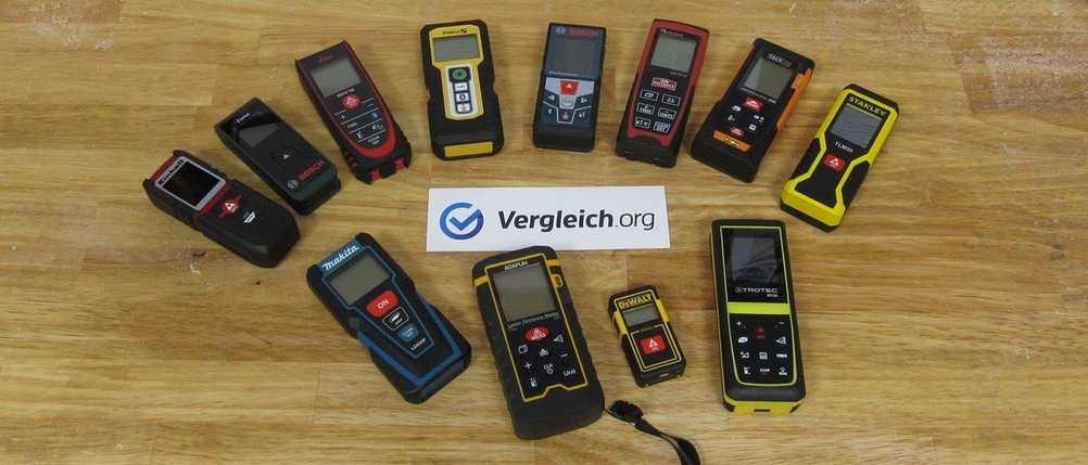 Einige Laserentfernungsmesser liegen mit einem Schild von Vergleich.org auf einem Tisch.