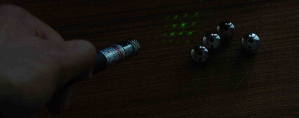 Hand hält einen getesteten Laserpointer, der ein grünes Licht-Muster auf eine dunkle Oberfläche, neben verschiedenen Lasermuster-Aufsätzen, wirft.