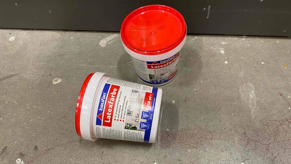 Zwei Eimer getestete Latexfarbe, einer liegend einer stehend auf einem Betonboden.