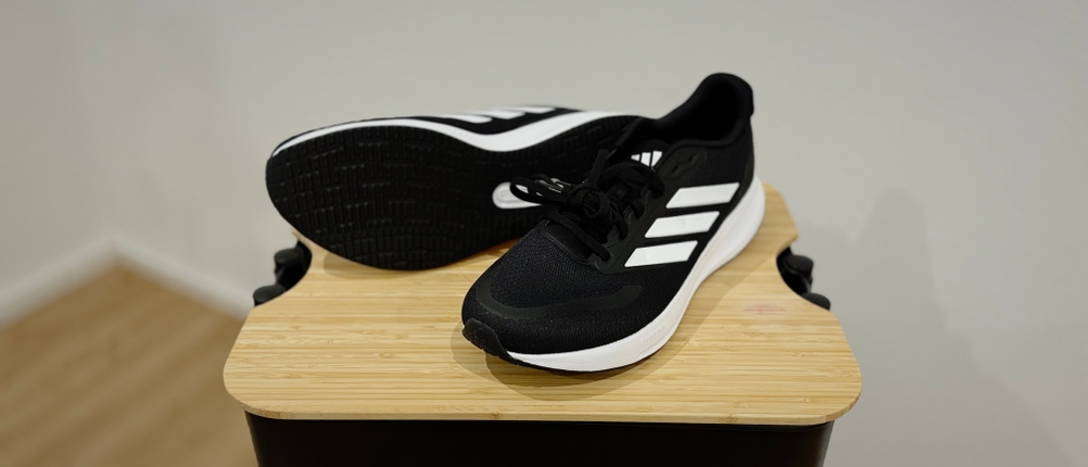 Schwarze Adidas Runfalcon 5 stehen auf einem Holzhocker. Einer der Schuhe, der rechte, liegt auf der Seite und gibt den Blick auf seine abgerundete schwarze Sohle frei.