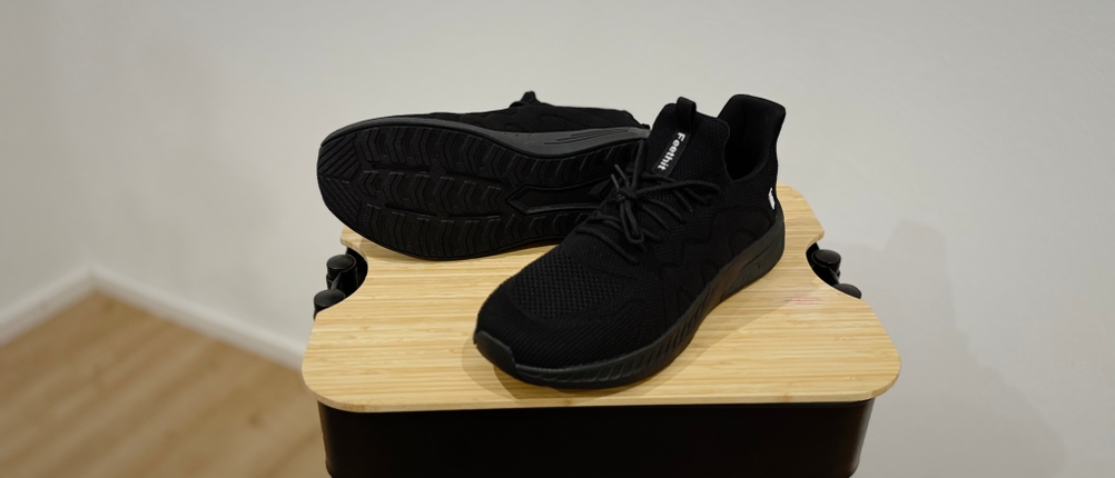 Feethit Sneaker Laufschuhe stehen auf einem Holzhocker. Einer der Schuhe, der rechte, liegt auf der Seite und gibt den Blick auf seine schwarze Sohle frei.