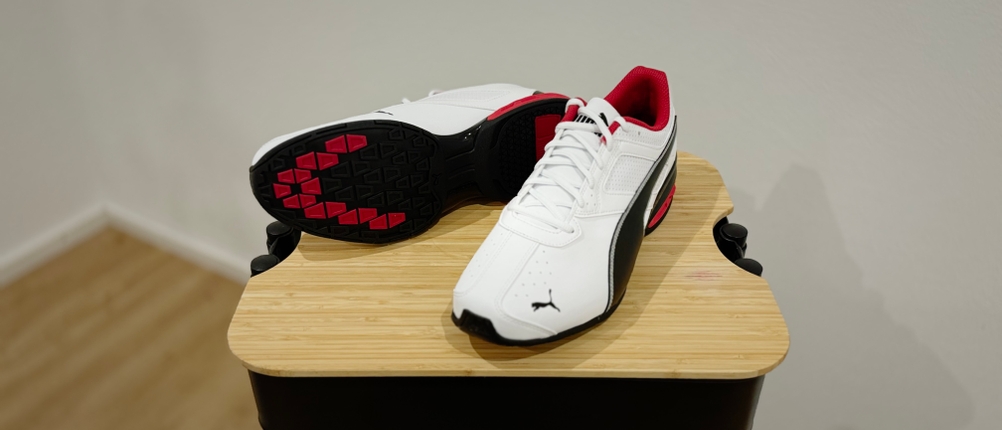 Weiße Puma Tazon 6 FM stehen auf einem Holzhocker. Einer der Schuhe, der rechte, liegt auf der Seite und gibt den Blick auf seine schwarz-rote Sohle frei.