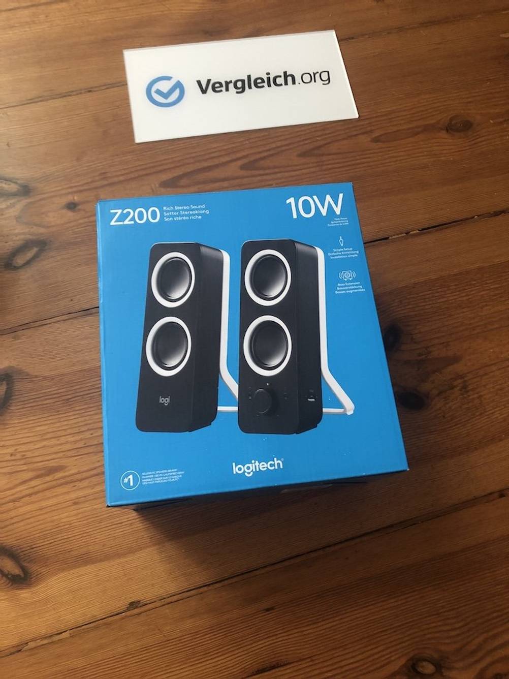 Logitech-Verpackung in Blau bei einem Lautsprecher-Test. Sie liegt mit einem Vergleich.org-Schild auf einer Holzoberfläche.