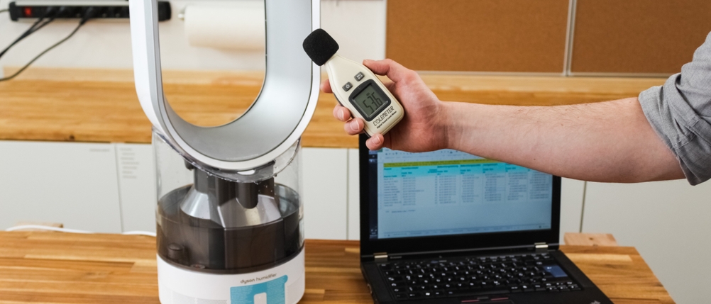 Eine Hand hält einen Geräuschmesser in der Hand vor einem Luftbefeuchter und Laptop.