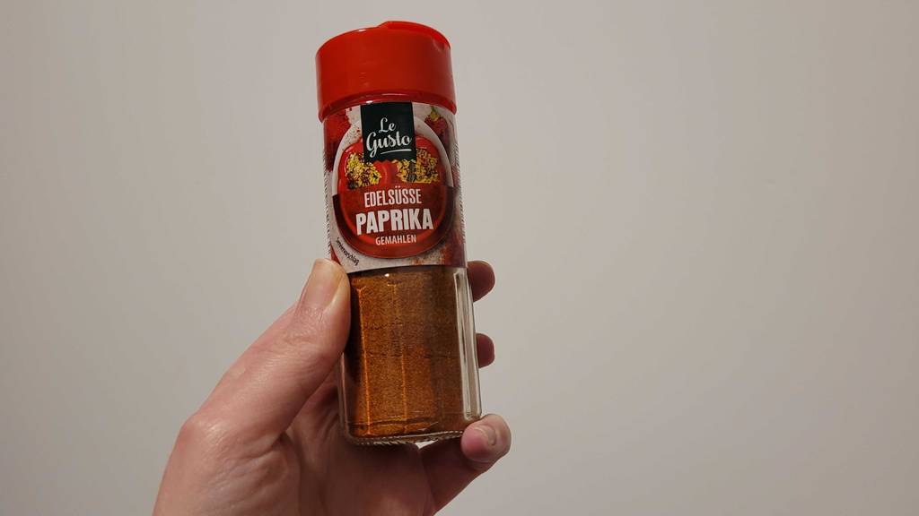 Eine Hand hält einen Gewürzstreuer mit getestetem Paprika-Edelsüß vor einer weißen Wand.