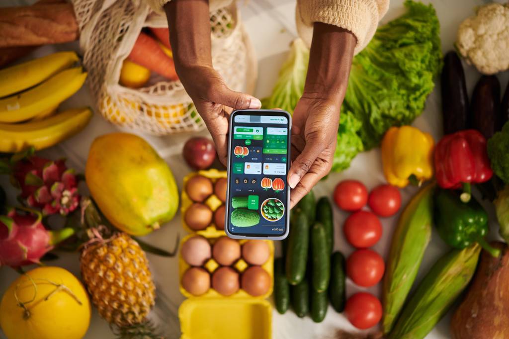 Lebensmittel online getestet: Eine Person hält ihr Smartphone mit einer Essensbestellung über einem Tisch voller Lebensmittel.