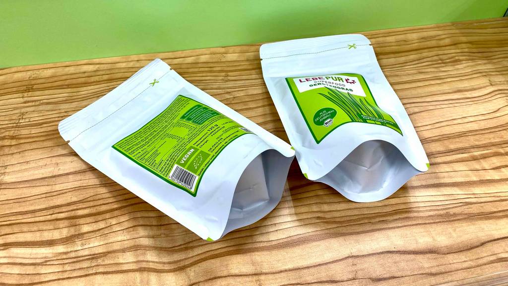 Zwei Standbeutel Lebepur Gerstengras im Test liegen mit Sicht auf die Vorder- und Rückseite inklusive Produktbeschreibung auf einer Holzfläche vor grüner Wand.