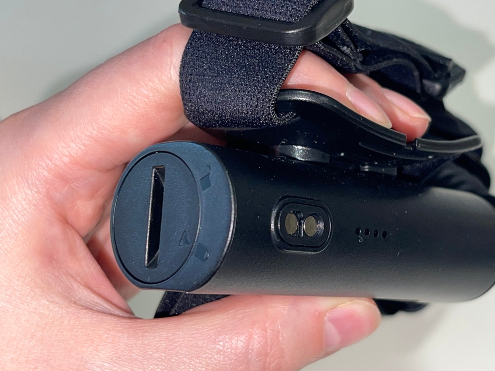 Der Akku der Stirnlampe Ledlenser H7R Core wird in einer Hand gehalten, gut sichtbar ist der Verschlussdeckel mit Bajonettverschluss. Auch die magnetische Ladebuchse und LED-Ladestandanzeige sind zu erkennen.