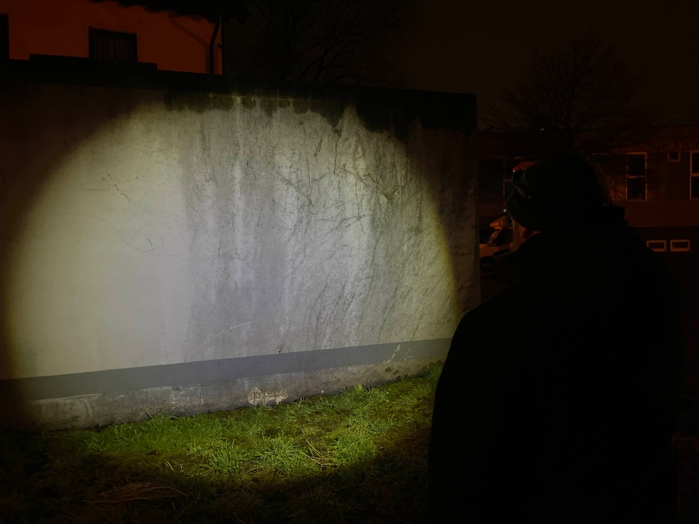 Ein Mann steht mit einer Ledlenser H7R Core Stirnlampe vor einer Wand. Die Lampe erzeugt einen großen Lichtkegel mit breitem Streuungsfeld.