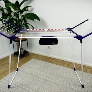 Der Leifheit-Standtrockner 81510 steht vollständig aufgeklappt in einem Raum auf einem Teppich. Das Gestell ist aus weißem Metall mit violetten Kunststoffelementen.