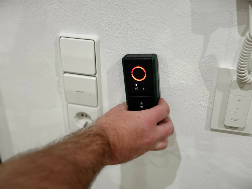 Eine Hand hält den Leitungssucher von Bosch an eine Wand neben einen Lichtschalter.