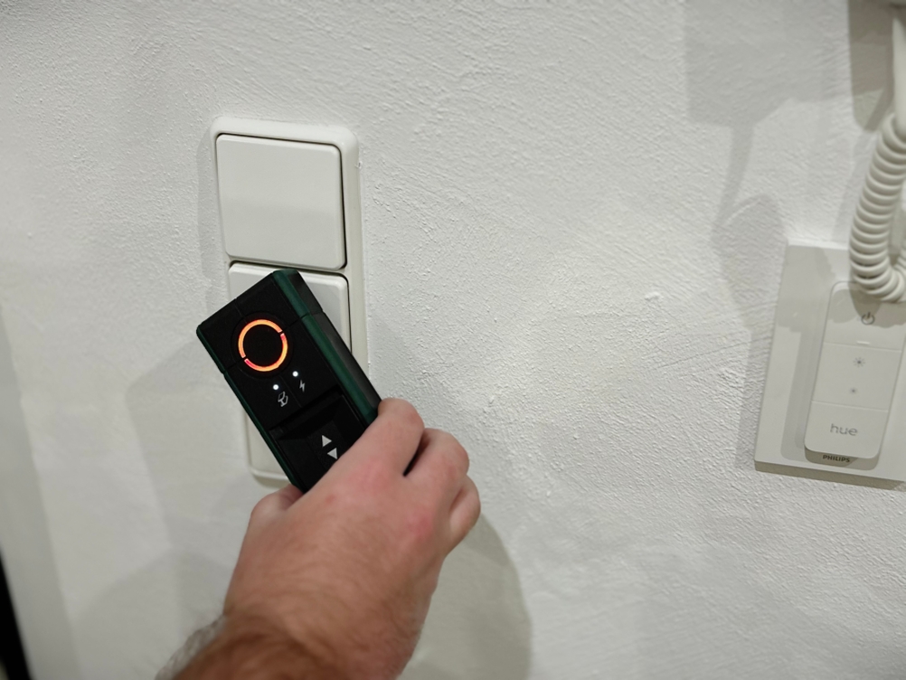 Eine Hand hält den Leitungssucher von Bosch an eine Wand vor einen Lichtschalter.