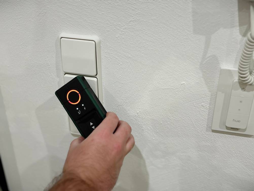 Eine Hand hält den Leitungssucher von Bosch an eine Wand vor einen Lichtschalter.