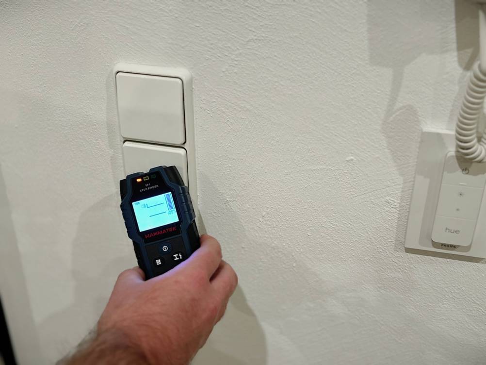 Eine Hand hält den Leitungssucher von Hanmatek an eine Wand vor einen Lichtschalter.