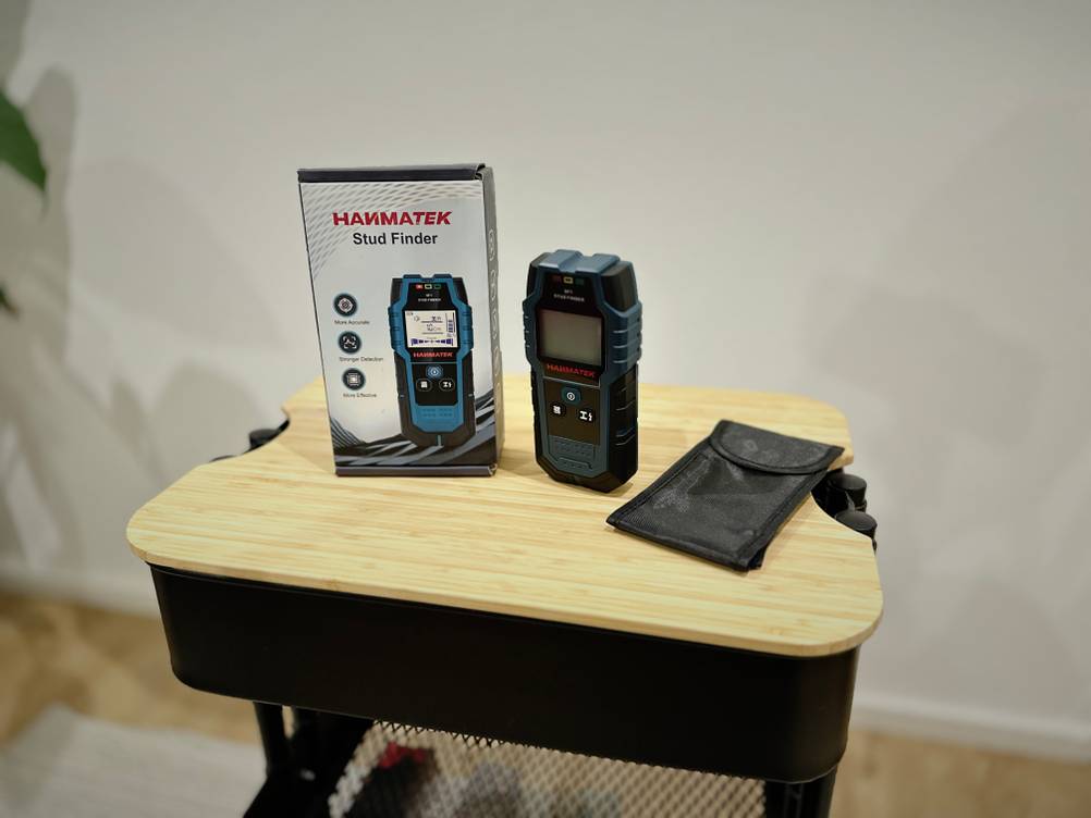 Das Produkt von Hanmatek steht mit der Verpackung und Zubehör auf einem hölzernen Untergrund vor einer weißen Wand.