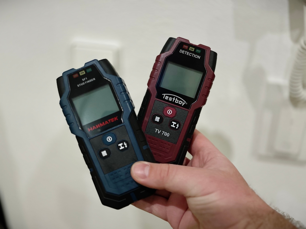 Zwei Leitungssucher werden mit einer Hand vor einer weißen Wand gehalten.