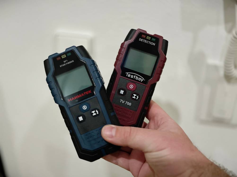 Zwei Leitungssucher werden mit einer Hand vor einer weißen Wand gehalten.