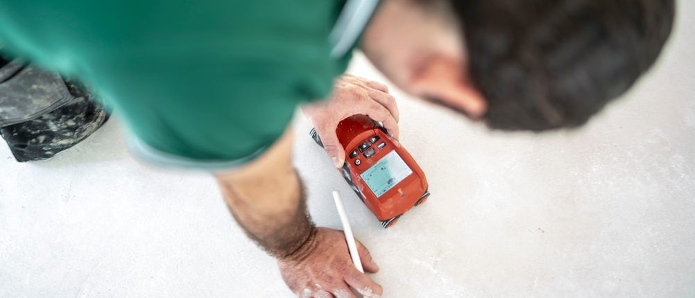 Mit einem Detektor ortet der Bodenleger die unsichtbaren Rohre der Fußbodenheizung, um anschließend eine präzise Messung der Estrichfeuchte durchführen zu können.