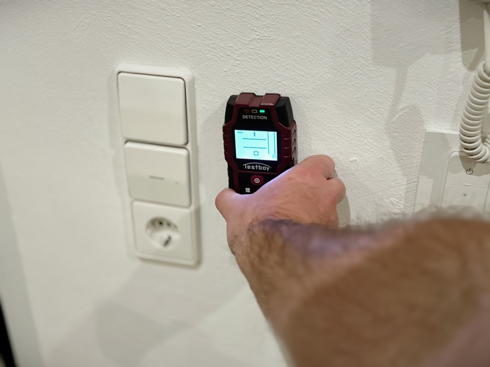 Eine Hand hält den Leitungssucher von Testboy an eine Wand neben einen Lichtschalter.